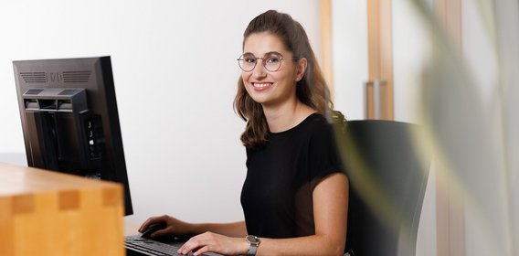 Die junge Wirtschaftsfachwirtin sitzt im Büro vor einem Computer und lächelt in die Kamera