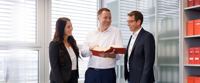 Drei Steuerberater stehen in den Büroräumen der Steuerkanzlei Nürnberg und schauen lächelnd in ein aufgeschlagenes Buch 