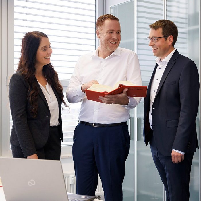 Drei Steuerberater stehen in den Büroräumen der Steuerkanzlei Nürnberg und schauen lächelnd in ein aufgeschlagenes Buch 